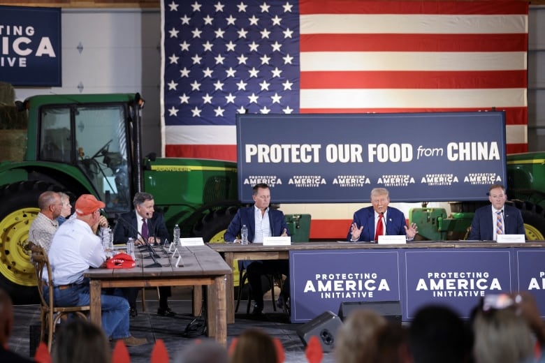 Trump at a table, in front of a green John Deere tractor and a sign "Protecting our food from China." 